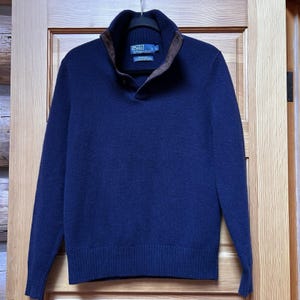 May include: A navy blue sweater with a brown collar and a buttoned neckline. The sweater has a ribbed hem and cuffs. The Polo by Ralph Lauren logo is visible on the inside collar.