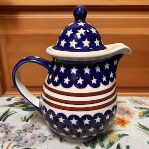 May include: A ceramic pitcher with a lid, featuring a blue and white star pattern on the lid and body. The body also has brown stripes. The pitcher has a curved handle and a spout for pouring.