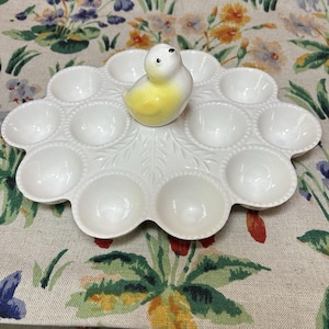 May include: White ceramic egg serving platter with 12 egg cups, shaped like a flower. A small, yellow and white bird figurine sits in the center. The platter has a leaf design and beaded edges. A decorative piece for serving eggs.