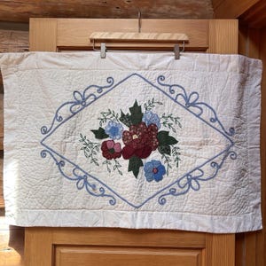 May include: A rectangular white quilted fabric piece featuring a central floral appliqué design with red, burgundy, and blue fabric flowers and green leaves. A light blue embroidered diamond-shaped border with decorative scrollwork surrounds the flowers.