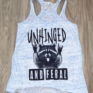 Puede incluir: Camiseta sin mangas azul claro con efecto jaspeado. La parte delantera presenta un gr&aacute;fico negro de un mapache haciendo un gesto de rock and roll, con las palabras "UNHINGED" arriba y "AND FERAL" abajo.