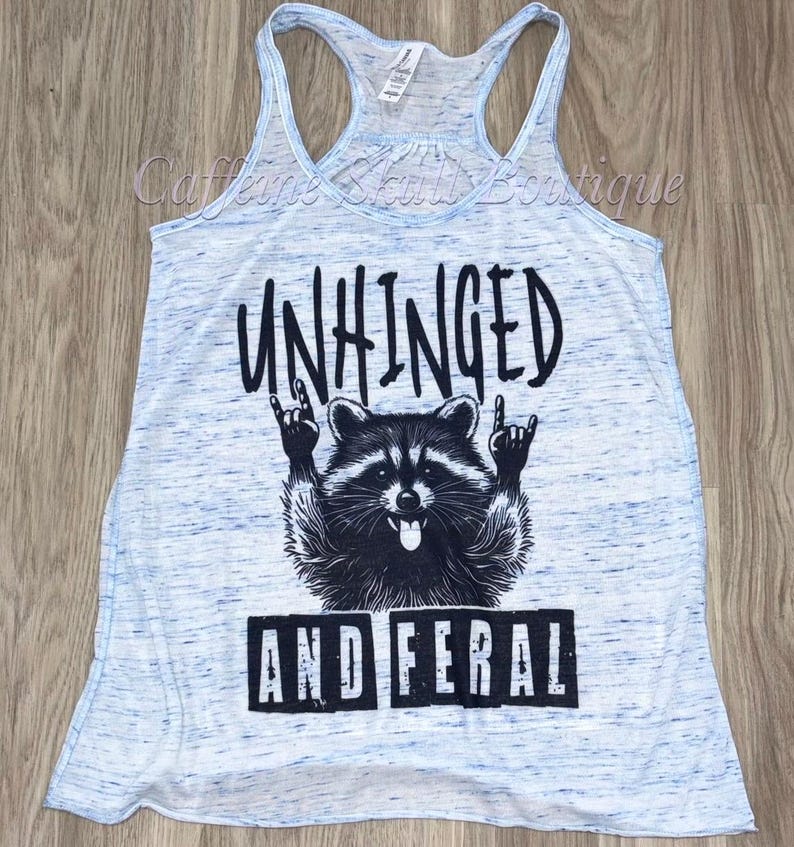 Puede incluir: Camiseta sin mangas azul claro con efecto jaspeado. La parte delantera presenta un gr&aacute;fico negro de un mapache haciendo un gesto de rock and roll, con las palabras "UNHINGED" arriba y "AND FERAL" abajo.