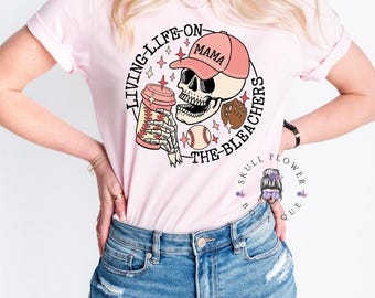 May include: Pink t-shirt with a graphic of a skull wearing a baseball cap that says "Mama" and holding a coffee cup and a baseball glove. The text around the skull says "Living Life On The Bleachers".