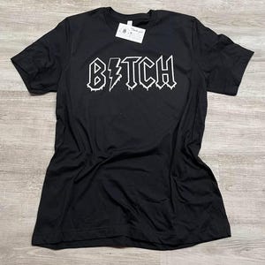 Pode incluir: Camiseta preta com um gr&aacute;fico branco da palavra "BITCH" em uma fonte de metal que pinga com um raio.