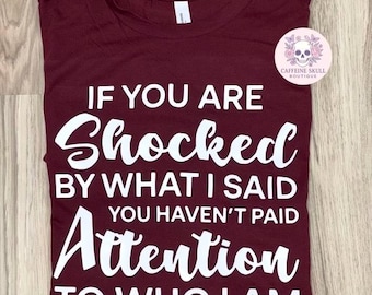 May include: A maroon short-sleeved t-shirt featuring white text that reads: IF YOU ARE Shocked BY WHAT I SAID YOU HAVEN'T PAID Attention TO WHO I AM. A circular logo with a skull and floral design labeled CAFFEINE SKULL BOUTIQUE is attached.