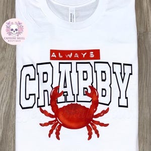 May include: White t-shirt with the words "ALWAYS CRABBY" in black block letters with a red crab graphic. The word "ALWAYS" is in a red rectangle above the word "CRABBY".