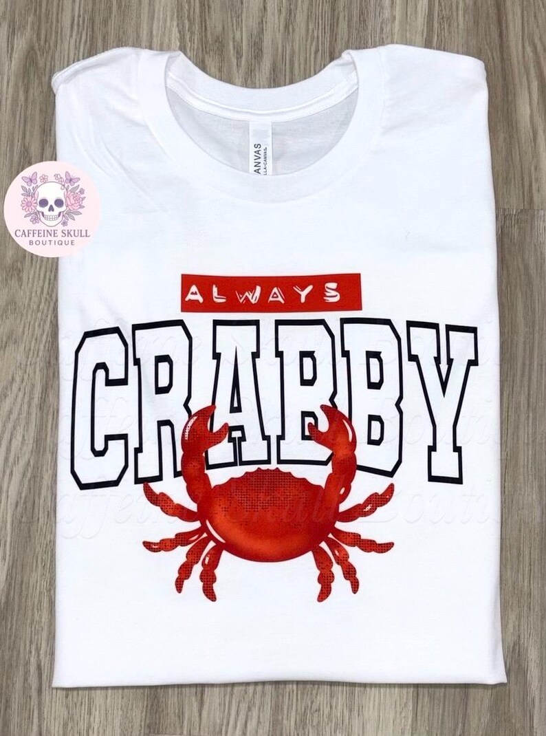 May include: White t-shirt with the words "ALWAYS CRABBY" in black block letters with a red crab graphic. The word "ALWAYS" is in a red rectangle above the word "CRABBY".