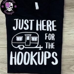 Puede incluir: Una camiseta negra de manga corta con el texto impreso en blanco JUST HERE FOR THE HOOKUPS y un gráfico de una caravana vintage. Un logotipo circular en la esquina superior izquierda muestra una calavera con flores y el texto Skull Flower Boutique.