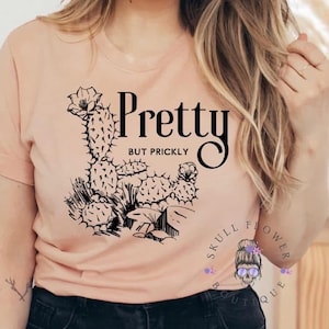 May include: A peach-colored t-shirt with a black graphic of a cactus and the words "Pretty but Prickly." The design includes a detailed illustration of a cactus with flowers and the text in a bold, stylized font.