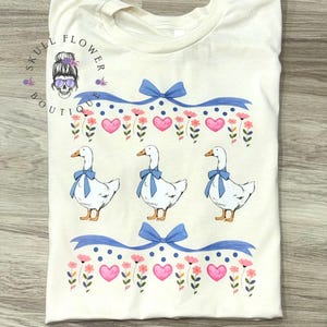以下が含まれることがあります： クリーム色のTシャツ。青い蝶ネクタイをした白いガチョウが3羽描かれています。ピンクのハート、花、青いリボンがデザインされています。「SKULL FLOWER BOUTIQUE」の文字が左上にプリントされています。