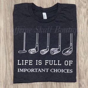 Puede incluir: Una camiseta de cuello redondo en color gris carbón oscuro con un gráfico blanco de cinco palos de golf diferentes alineados sobre el texto blanco que dice: LIFE IS FULL OF IMPORTANT CHOICES. La camiseta tiene mangas cortas.