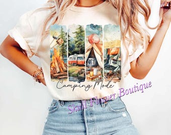 Puede incluir: Una camiseta blanca con una ilustración de acuarela de una escena de camping. La ilustración incluye una fogata, una tienda de campaña, una camioneta y una mesa de picnic. El texto "Camping Mode" está escrito en cursiva negra debajo de la ilustración. El texto "Skull Flower Boutique" está escrito en cursiva morada debajo del texto "Camping Mode".