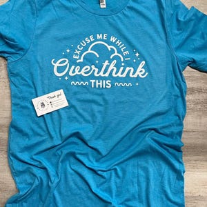 May include: A turquoise t-shirt with the phrase "EXCUSE ME WHILE I OVERTHINK THIS" in white script and a cloud graphic. The shirt is made of soft material and has a crew neck. A small card is visible near the bottom.