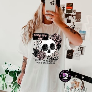 以下が含まれることがあります： 「may we all heal from the things we don't talk about」の文字と、スカルとバラのデザインが施された白いTシャツ。デザインにはスカル、バラ、星が含まれています。シャツには「Skull Flower Boutique」と書かれたロゴも付いています。