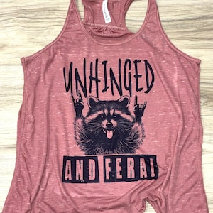 Puede incluir: Una camiseta sin mangas rosa polvorienta con un gr&aacute;fico de un mapache haciendo un gesto de rock and roll con la mano. El texto en la camiseta dice "UNHINGED" encima del mapache y "AND FERAL" debajo.