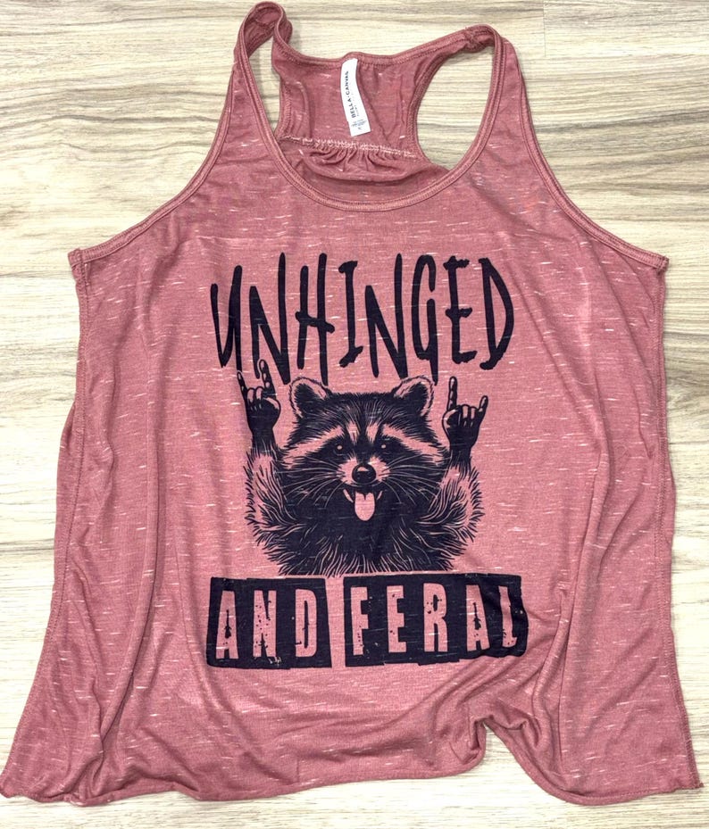 Puede incluir: Una camiseta sin mangas rosa polvorienta con un gr&aacute;fico de un mapache haciendo un gesto de rock and roll con la mano. El texto en la camiseta dice "UNHINGED" encima del mapache y "AND FERAL" debajo.