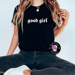 May include: Black t-shirt featuring the phrase "good girl" in white gothic lettering. The shirt is worn with dark denim jeans and white trainers. A circular logo with a skull and flowers is visible.
