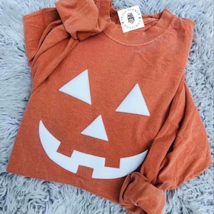 Könnte beinhalten: Orangefarbenes Langarmshirt mit einem weißen Kürbisgesicht-Design. Das Shirt hat einen Rundhalsausschnitt und hochgekrempelte Ärmel. Ein kleines Etikett mit Text ist in der Nähe des Halsausschnitts sichtbar. Das Shirt ist aus weichem Material.