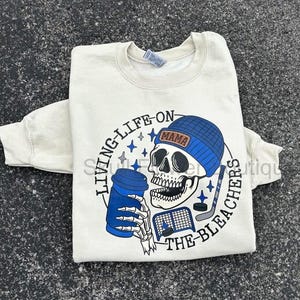 May include: Cream-coloured sweatshirt featuring a skull wearing a blue beanie with the word "MAMA". The skull holds a blue coffee cup and hockey stick. Text reads "LIVING-LIFE-ON-THE-BLEACHERS".