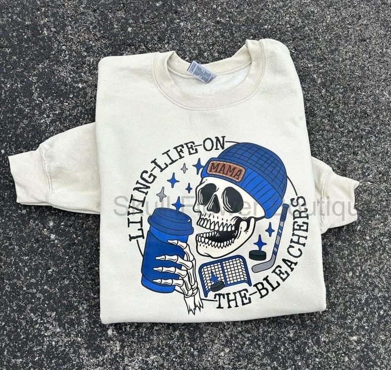 May include: Cream-coloured sweatshirt featuring a skull wearing a blue beanie with the word "MAMA". The skull holds a blue coffee cup and hockey stick. Text reads "LIVING-LIFE-ON-THE-BLEACHERS".