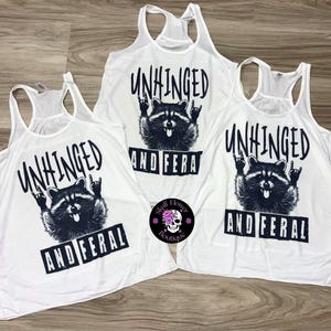 Puede incluir: Camisetas sin mangas blancas con un gr&aacute;fico negro de un mapache haciendo un gesto de rock and roll. El texto "UNHINGED" y "AND FERAL" est&aacute; impreso encima y debajo de la ilustraci&oacute;n del mapache.