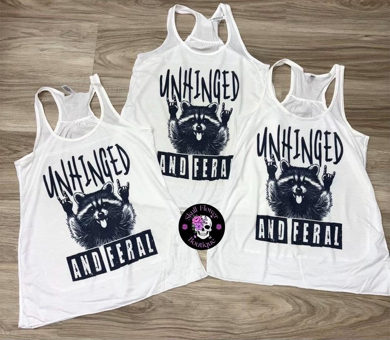Puede incluir: Camisetas sin mangas blancas con un gr&aacute;fico negro de un mapache haciendo un gesto de rock and roll. El texto "UNHINGED" y "AND FERAL" est&aacute; impreso encima y debajo de la ilustraci&oacute;n del mapache.