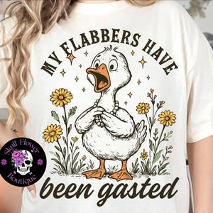 Könnte beinhalten: Cremefarbenes T-Shirt mit einem Cartoon-Enten-Motiv, das eine Perlenkette trägt und von Blumen umgeben ist. Der Text "MY FLABBERS HAVE been gasted" ist über und unter der Ente aufgedruckt. Ein Skull Flower Boutique-Logo befindet sich unten links.