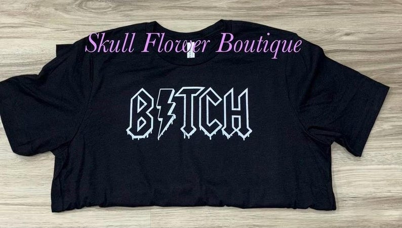 Pode incluir: Camiseta preta com um gr&aacute;fico branco que diz "B*TCH" em uma fonte g&oacute;tica que pinga.