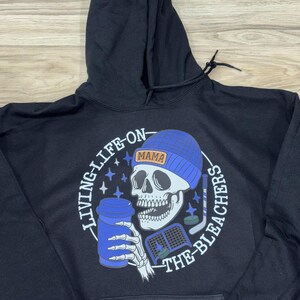 May include: Black hoodie featuring a skull wearing a blue beanie with the word "MAMA". The skull holds a blue coffee cup and hockey stick. The text "LIVING-LIFE-ON-THE-BLEACHERS" is printed around the graphic.