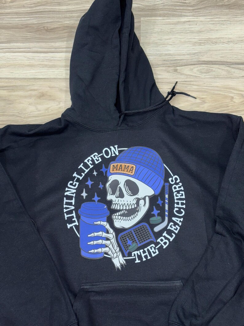 May include: Black hoodie featuring a skull wearing a blue beanie with the word "MAMA". The skull holds a blue coffee cup and hockey stick. The text "LIVING-LIFE-ON-THE-BLEACHERS" is printed around the graphic.