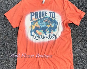May include: Orange t-shirt with a white bleached design of a buffalo silhouette with mountains and trees in the background. The text "Prone to Wander" is printed below the buffalo. The text "Skull Flower Boutique" is printed at the bottom of the shirt.