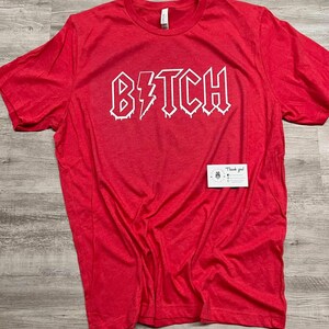 Pode incluir: Camiseta vermelha com um gr&aacute;fico branco que diz "B*TCH" em uma fonte de rock and roll que pinga.