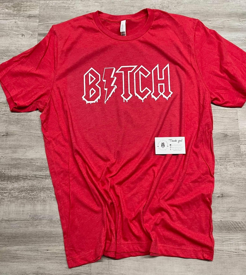 Pode incluir: Camiseta vermelha com um gr&aacute;fico branco que diz "B*TCH" em uma fonte de rock and roll que pinga.