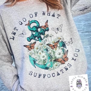 Puede incluir: Sudadera gris con el texto "Let go of what suffocates you" y un ancla turquesa con flores y mariposas.