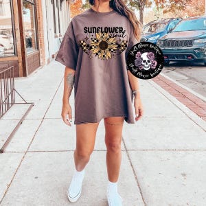 Puede incluir: Una camiseta marrón de gran tamaño con el texto "Sunflower Soul" y un gráfico de girasol con un diseño de bandera de carreras a cuadros. La camiseta la lleva puesta una persona que está de pie en una acera.