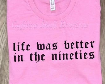 Könnte beinhalten: Ein pinkfarbenes T-Shirt mit dem Text "life was better in the nineties" in schwarzer gotischer Schrift. Das T-Shirt zeigt auch den Text "Caffeine Skull Boutique". Das T-Shirt ist auf einer Holzoberfläche gefaltet.