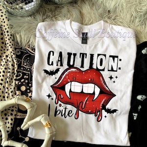 Puede incluir: Camiseta blanca con un gráfico de labios de vampiro rojos, goteando sangre, y el texto "CAUTION: I bite." El diseño incluye murciélagos y estrellas. La camiseta es de Caffeine Skull Boutique.