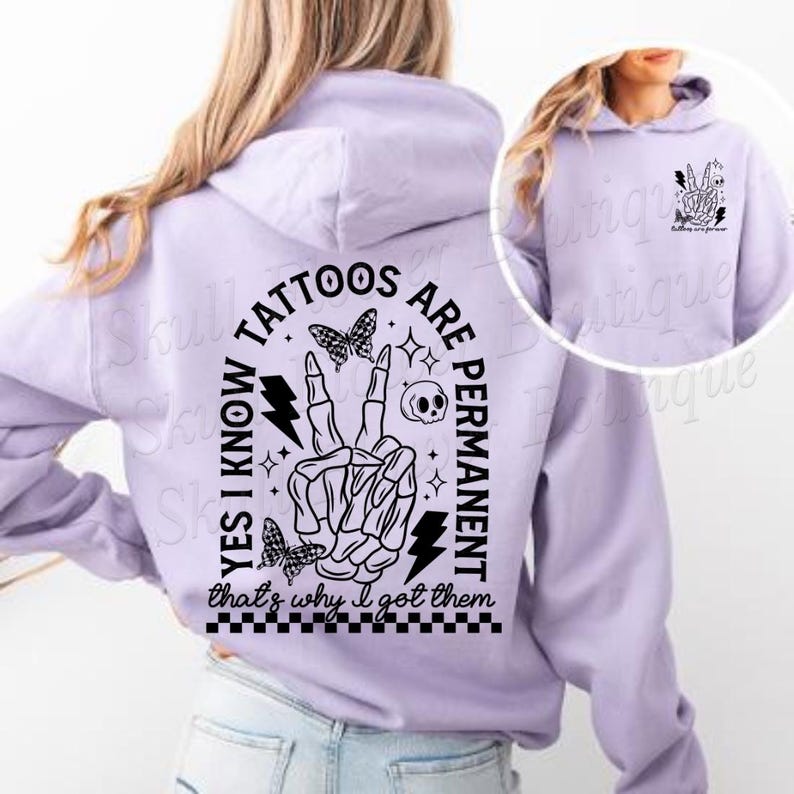 Op de afbeelding: Een lavendelkleurige hoodie met een zwarte afbeelding op de rug die zegt "Ja, ik weet dat tatoeages permanent zijn, daarom heb ik ze laten zetten". De afbeelding bevat een skelet hand die een vredesteken maakt, bliksem, vlinders en een schedel.
