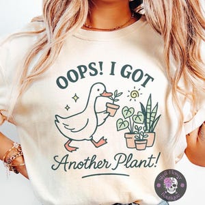 Pode incluir: Camiseta creme com um gráfico extravagante. O design apresenta um pato de desenho animado carregando uma planta em vaso, acompanhado do texto "OOPS! I GOT Another Plant!" e plantas em vaso adicionais. O logotipo da Skull Flower Boutique está no canto inferior direito.