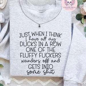 Puede incluir: Una sudadera gris claro con el texto "JUST WHEN I THINK I have all my DUCKS IN A ROW ONE OF THE FLUFFY FUCKERS wanders off and GETS INTO some shit" en negro. Se ve un pequeño collar con colgante.