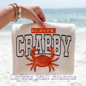 May include: Cream-colored beach towel with the words "ALWAYS CRABBY" in black block letters and a red crab graphic. The towel is held up by a hand with a bracelet. The background is a beach scene with the ocean.