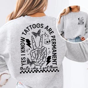 Op de afbeelding: Grijs sweatshirt met een zwart grafisch ontwerp met de tekst "Ja, ik weet dat tatoeages permanent zijn, daarom heb ik ze laten zetten". De grafiek bevat een skelet hand die een vredesteken maakt, een bliksem, een schedel en vlinders.