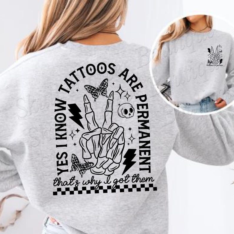 Op de afbeelding: Grijs sweatshirt met een zwart grafisch ontwerp met de tekst "Ja, ik weet dat tatoeages permanent zijn, daarom heb ik ze laten zetten". De grafiek bevat een skelet hand die een vredesteken maakt, een bliksem, een schedel en vlinders.