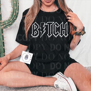 Pode incluir: Uma camiseta preta com a palavra "B*tch" escrita em branco com um raio e um efeito de gotejamento.