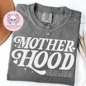 Puede incluir: Una camiseta gris con la palabra "MOTHERHOOD" en letras blancas grandes. La camiseta también tiene el texto "SOME DAYS I ROCK IT. SOME DAYS IT ROCKS ME. EITHER WAY WE'RE ROCKIN'". Un collar dorado y un sombrero también están en la imagen.