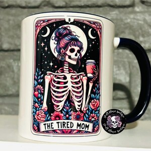 May include: White ceramic mug with a blue handle. The mug features a colorful illustration of a skeleton with a coffee cup, surrounded by flowers and stars. The text "The Tired Mom" is printed on the mug.
