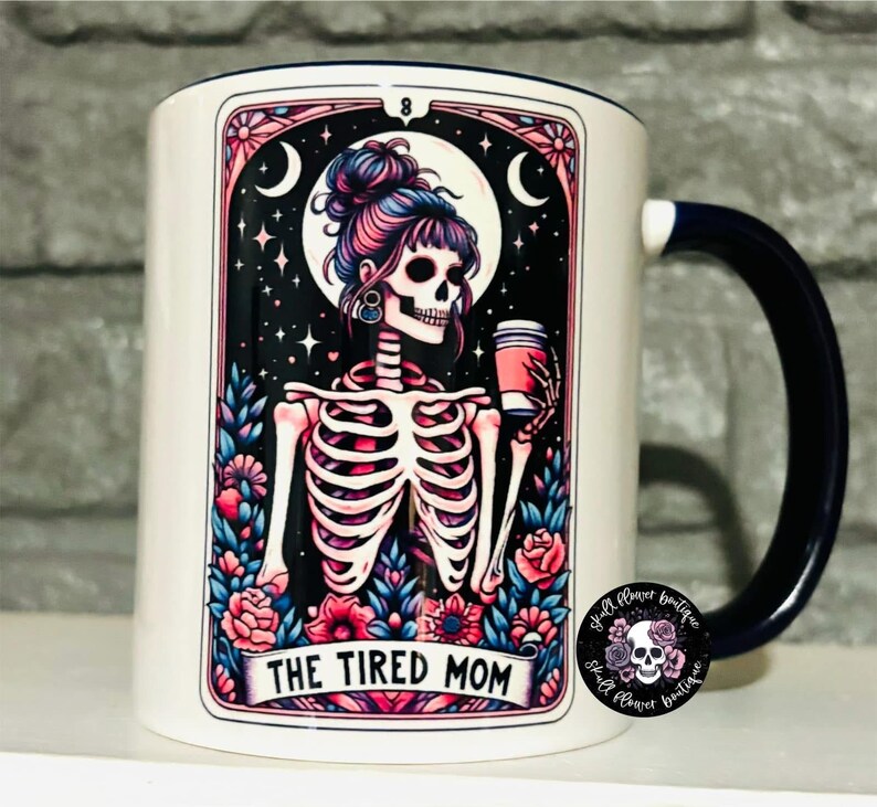 May include: White ceramic mug with a blue handle. The mug features a colorful illustration of a skeleton with a coffee cup, surrounded by flowers and stars. The text "The Tired Mom" is printed on the mug.