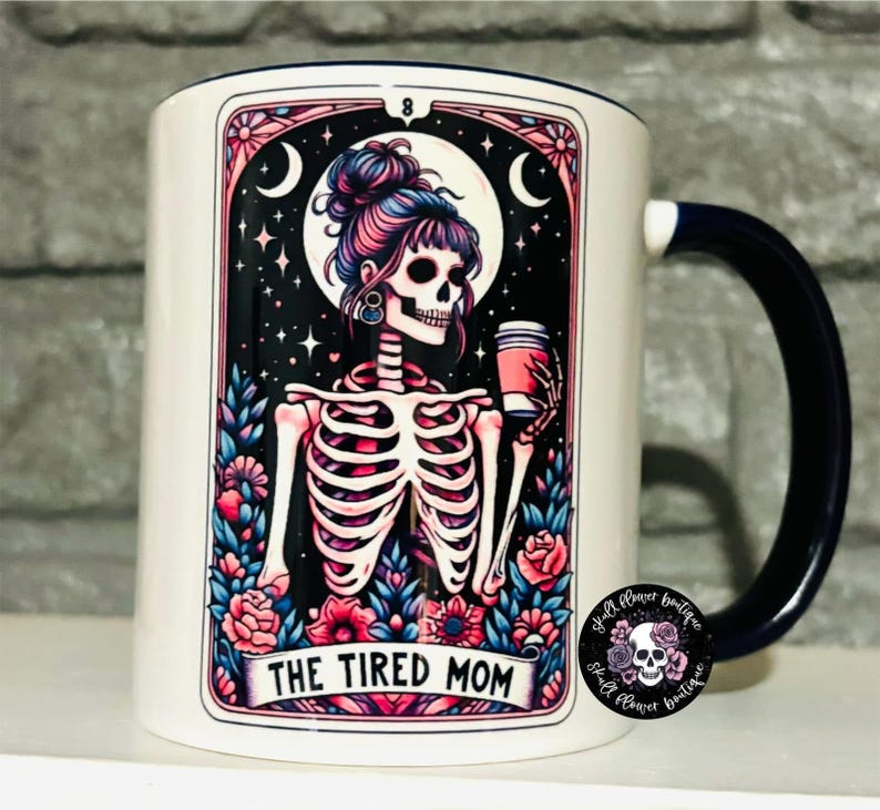May include: White ceramic mug with a blue handle. The mug features a colorful illustration of a skeleton with a coffee cup, surrounded by flowers and stars. The text "The Tired Mom" is printed on the mug.