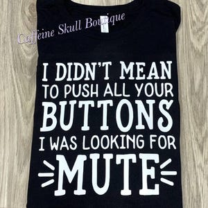 Op de afbeelding: Zwart T-shirt met de witte tekst: "I DIDN'T MEAN TO PUSH ALL YOUR BUTTONS I WAS LOOKING FOR MUTE". De merknaam "Caffeine Skull Boutique" staat in cursief bovenaan. Het T-shirt is opgevouwen.