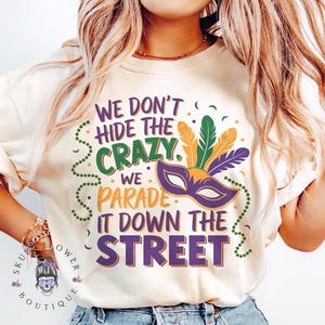 Może przedstawiać: Biała koszulka z nadrukiem w kolorze fioletowym, zielonym i złotym nawiązującym do Mardi Gras. Na koszulce widnieje napis "We don't hide the crazy, we parade it down the street".